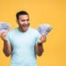 Trading Lab Academy - winner young rich happy african american indian black man in casual holding money dollar bills with surprise isolated over yellow wall background